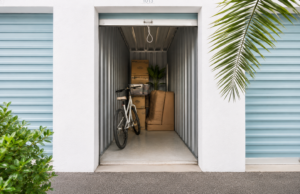 Open storage unit filled with a white bicycle, stacked cardboard boxes, a large brown sofa, and a potted plant inside.