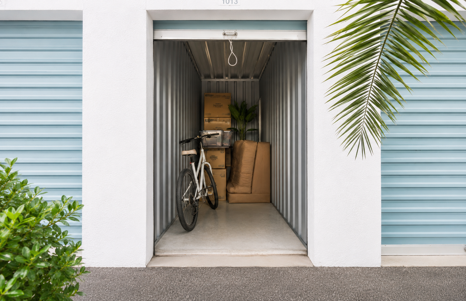 Open storage unit filled with a white bicycle, stacked cardboard boxes, a large brown sofa, and a potted plant inside.